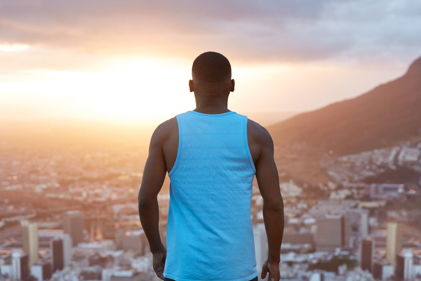 Sporty black male with strong muscular body, stands back, thinks about something while admires nature view, mountains and city from above, looks at sky with sunrise. Sportsman enjoys freedom